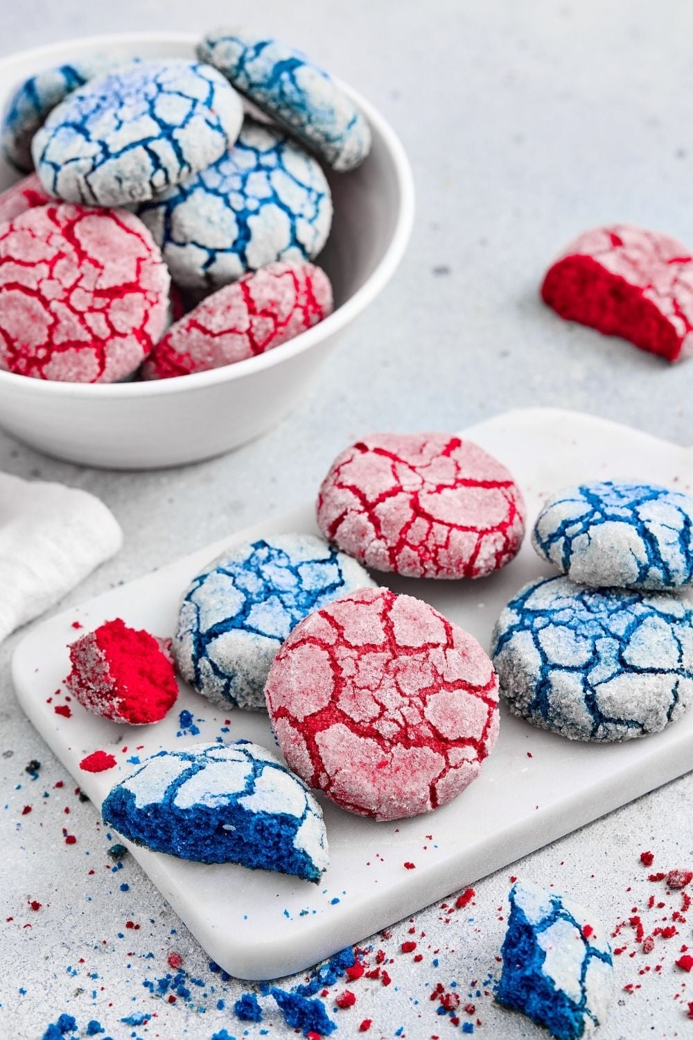 An angled shot showing multiple red and blue crinkle cookies, including two halved pieces revealing their vibrant interiors, displayed on a small marble board and scattered on a textured grey counter, with a white bowl of stacked cookies in the background, all sprinkled with colorful crumbs for a festive presentation.