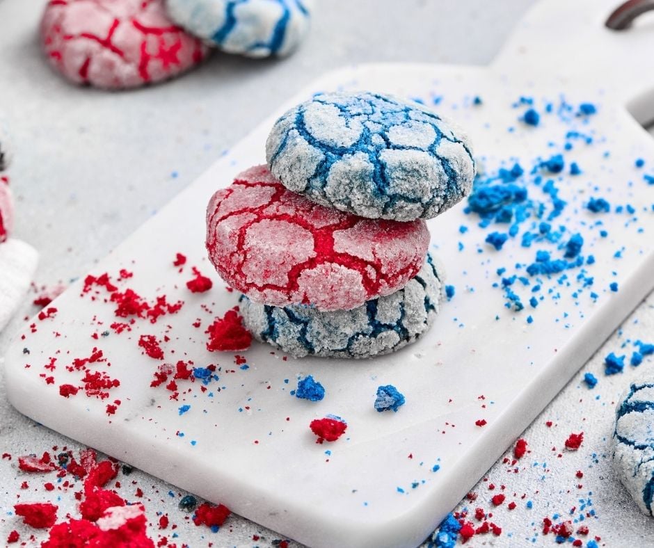 A three-quarter view of the finished red and blue crinkle cookies, focusing on a stack of two cookies—one bright red, one vibrant blue—sitting centrally on a white marble serving board. Small red and blue crumbs are scattered across the board, which has a distinct metal handle on the right. Additional cookies are visible, out of focus, in the upper-left and lower-right, all on a lightly textured grey counter.