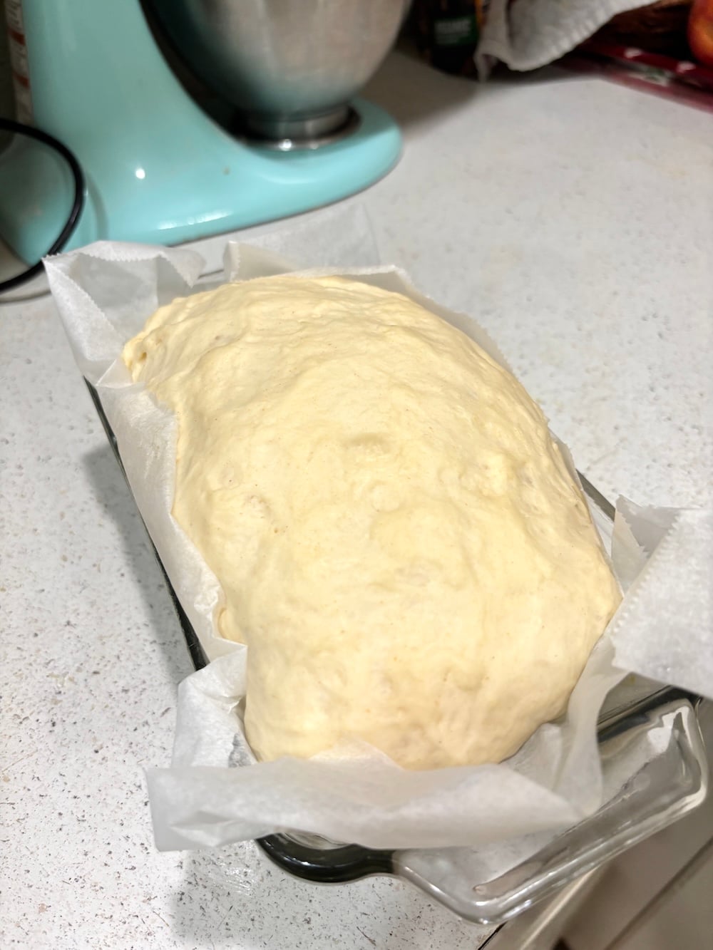 White bread dough in a glass loaf pan, risen above the rim.