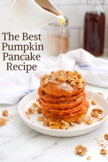 A stack of five pumpkin spice pancakes on a white plate, topped with chopped walnuts and drizzled with maple syrup. The background is a rustic wooden table with a napkin and other breakfast items, out of focus.